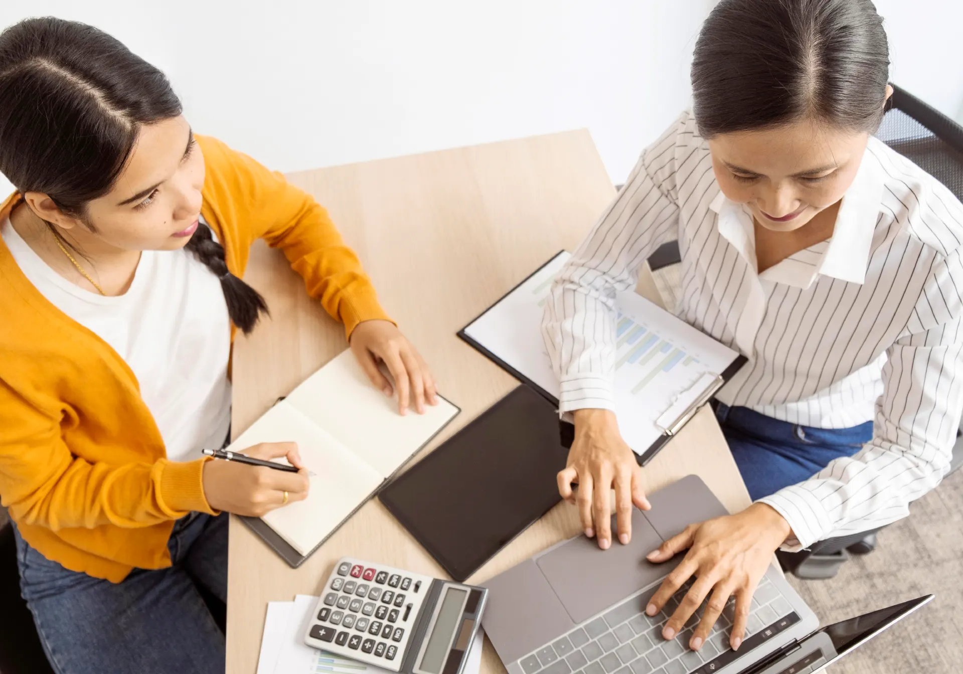 Dos mujeres trabajando juntas en un escritorio con un portátil, una libreta y una calculadora