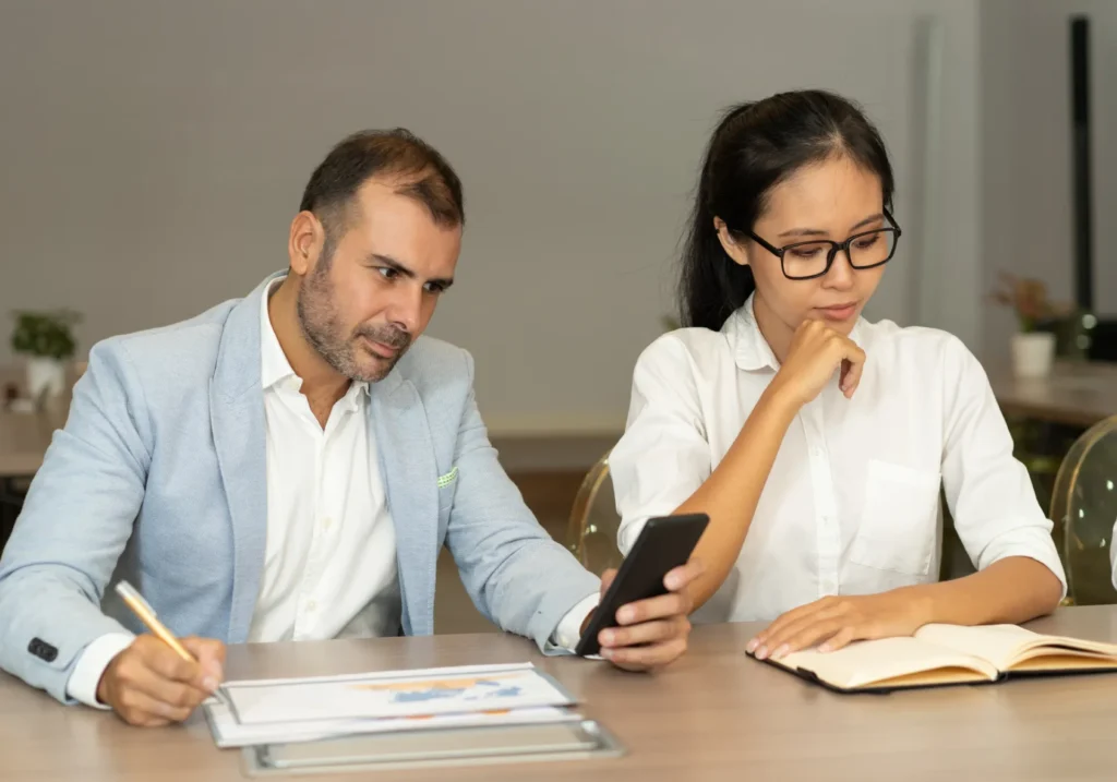 Un hombre y una mujer trabajando en un escritorio mientras el hombre mira el teléfono de su compañera